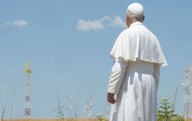 Pope Leo XIV stands in a field, gazing at communication towers against a blue sky.