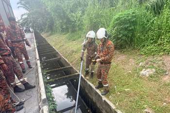 Two firefighters in camouflage uniforms examine a drainage ditch, using a long pole to investigate the water.