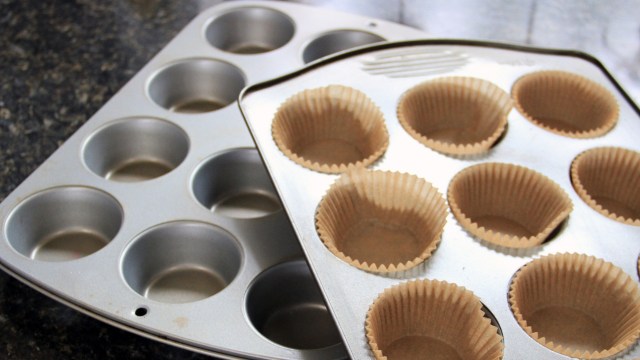 A close-up of two muffin trays with brown paper liners in one tray on a dark countertop.