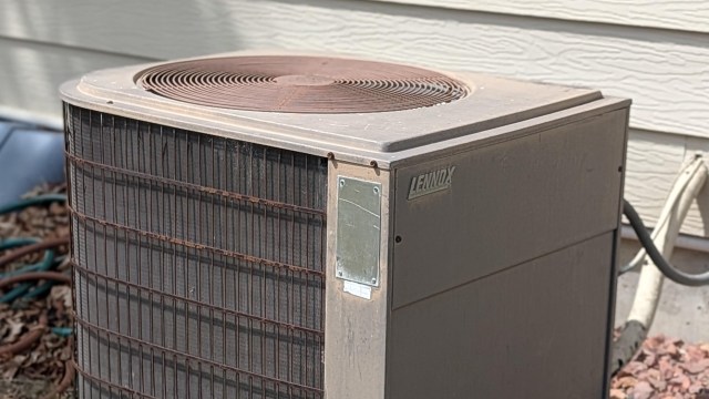 A weathered Lennox air conditioning unit sits beside a house, partially surrounded by landscaping.