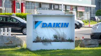A sign displaying the logo of Daikin, surrounded by grass and stones, with cars visible in the background.
