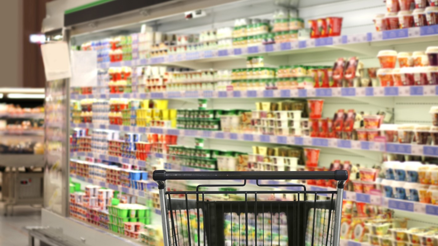 A shopping cart in front of colorful shelves filled with various dairy products in a supermarket.