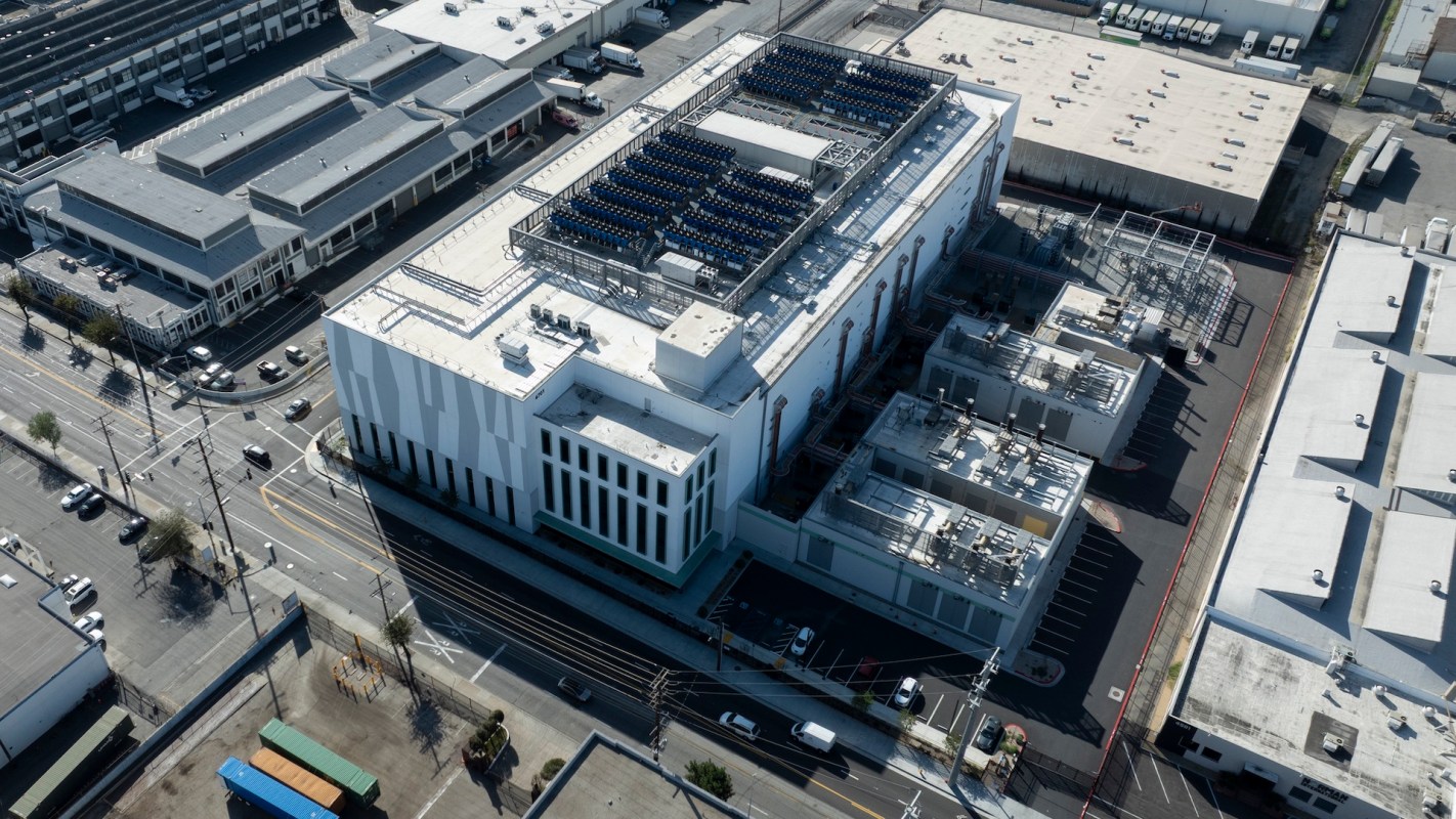 An aerial view of a modern industrial building with solar panels and surrounding vehicles on city streets.