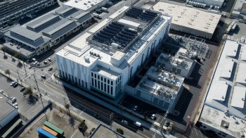 An aerial view of a modern industrial building with solar panels and surrounding vehicles on city streets.