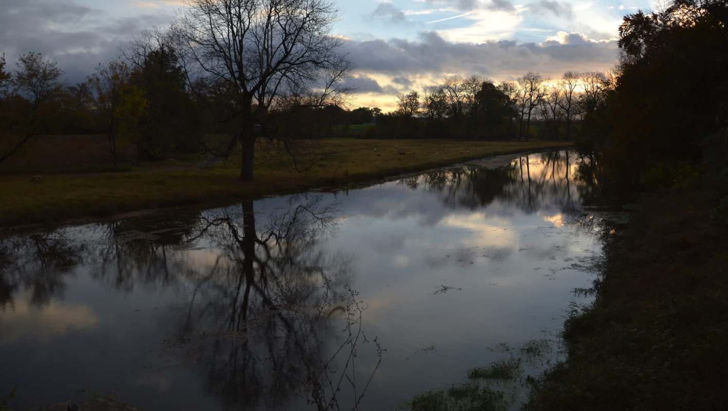 A tranquil riverside scene at dusk, reflecting trees and clouds under a moody sky.