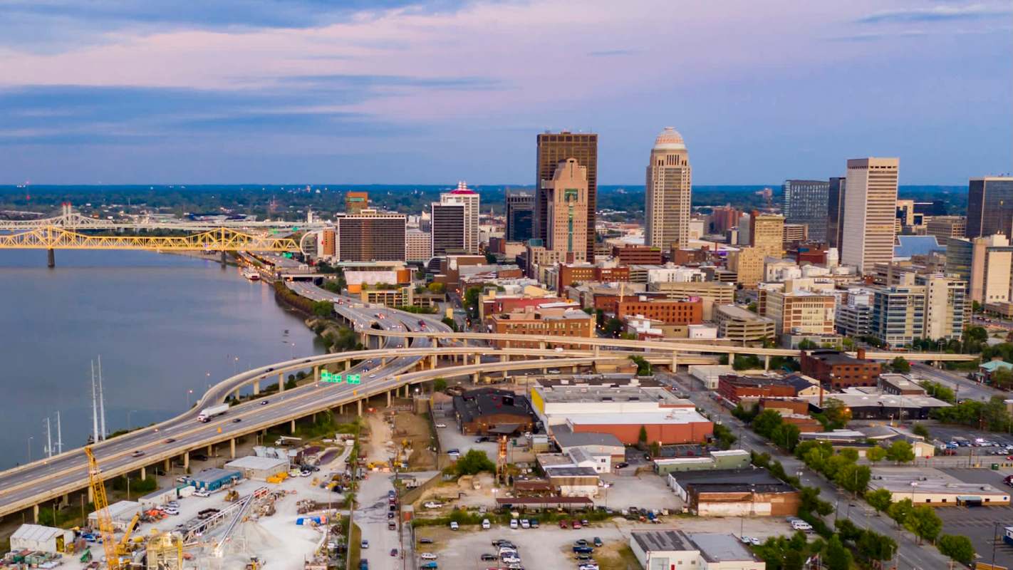 Aerial view of a city skyline with a river, highway, and modern buildings against a sunset sky.