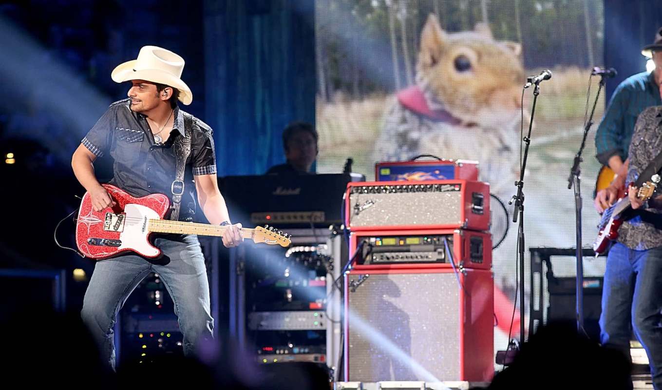 Country star Brad Paisley, in a cowboy hat, plays a red and white guitar on stage with a squirrel image displayed behind him.