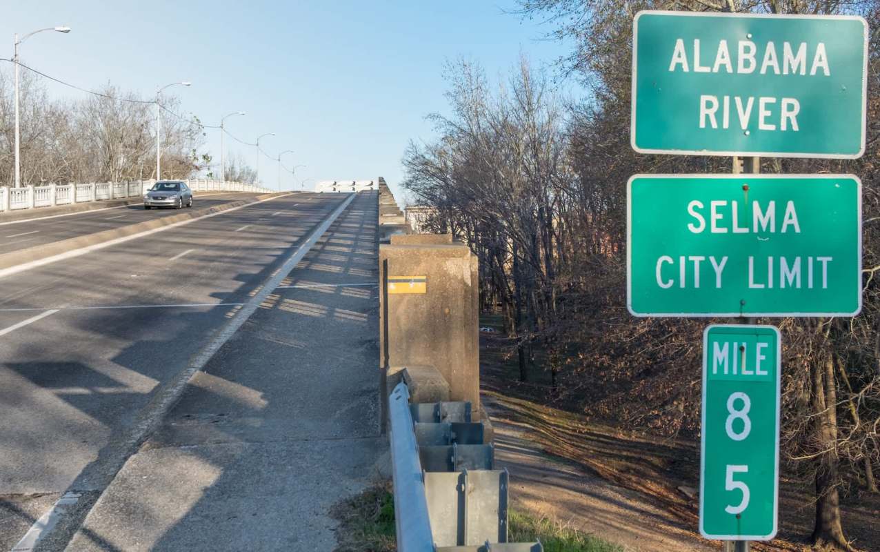 A highway sign indicating the Selma city limit and Alabama River, with a car driving up the road.