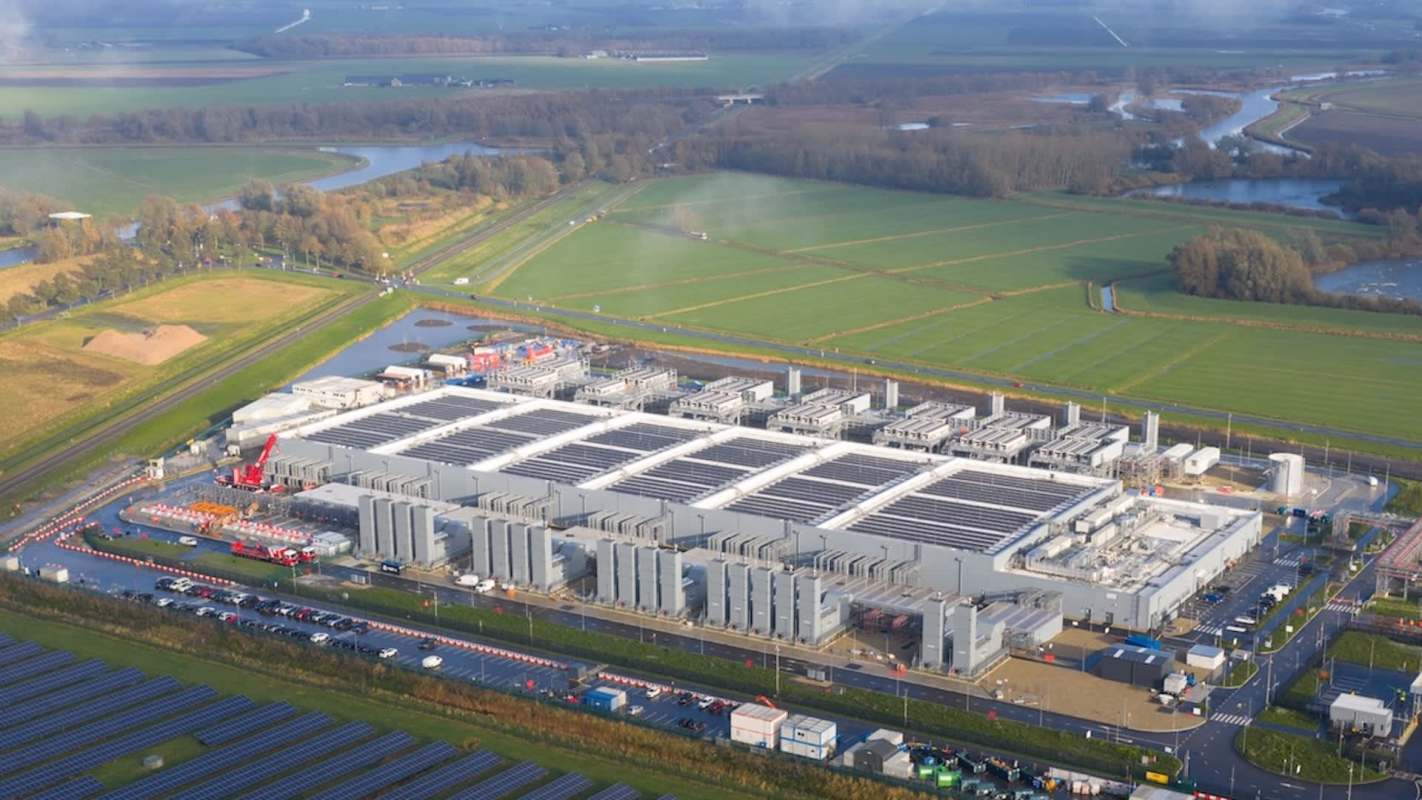 An aerial view of a solar panel field beside a large industrial facility in a rural landscape.