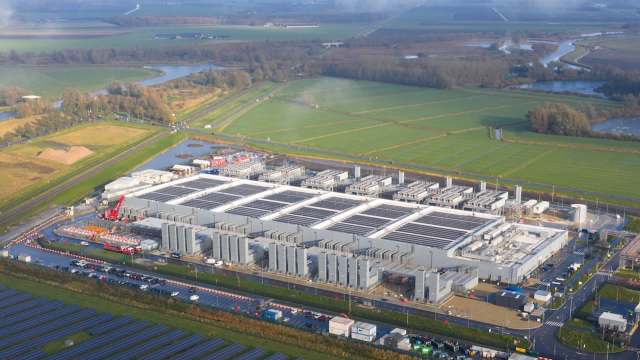 An aerial view of a solar panel field beside a large industrial facility in a rural landscape.