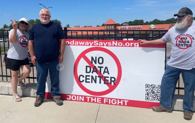 Three protestors stand beside an anti-data center sign.