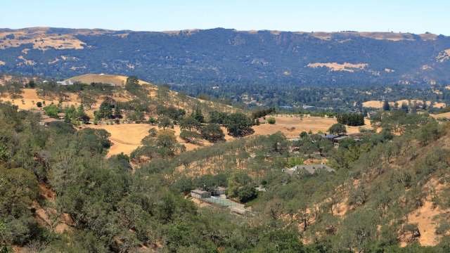 A panoramic view of rolling hills with scattered trees and residential areas under a clear blue sky.