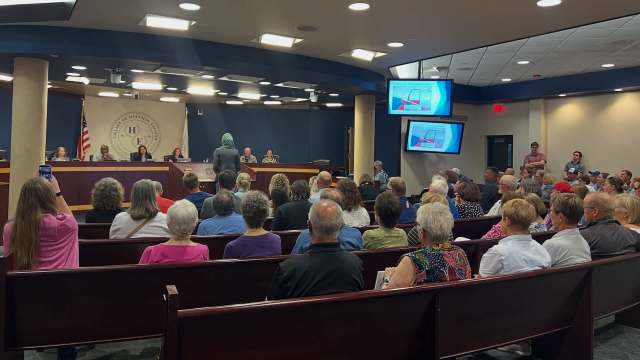 A crowded town hall meeting with audience members facing a speaker at a podium and screens displaying information.