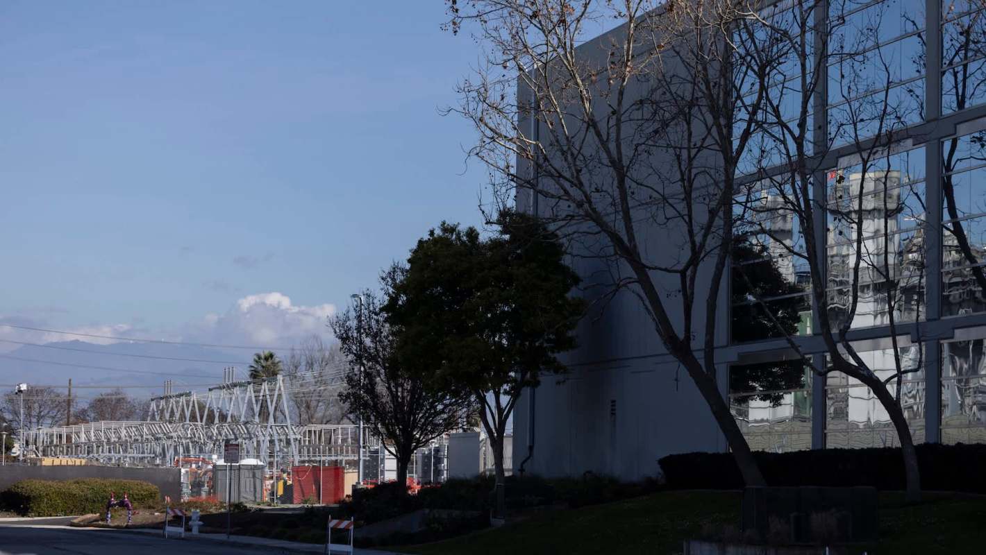 A modern building with reflective glass panels beside a power facility and several bare trees under a blue sky.