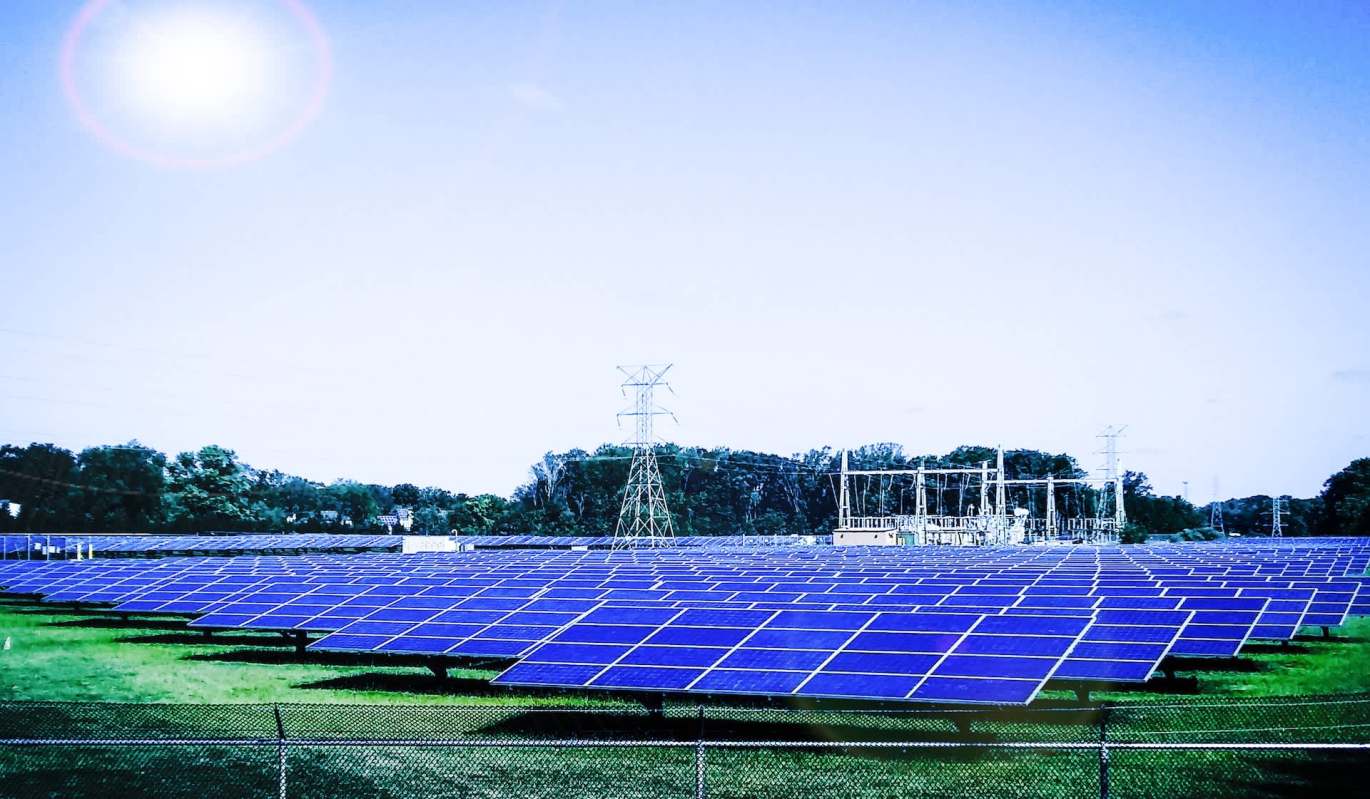 A large solar panel field under a bright sky, with power lines and equipment in the background.
