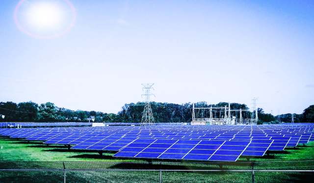A large solar panel field under a bright sky, with power lines and equipment in the background.