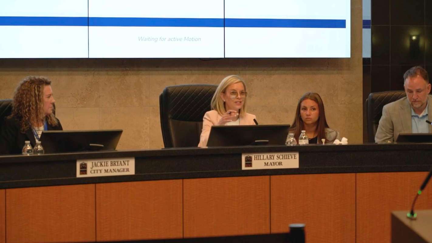 A mayor and city manager participate in a meeting at a city council table with screens in the background.
