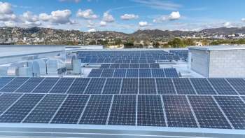 A rooftop covered in solar panels with a view of hills and buildings in the background under a partly cloudy sky.