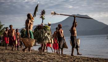 A group of men in traditional attire carry baskets and ceremonial objects along a beach with mountains in the background.