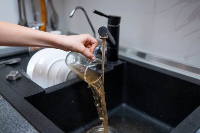 A person pours dirty water from a glass into a kitchen sink.