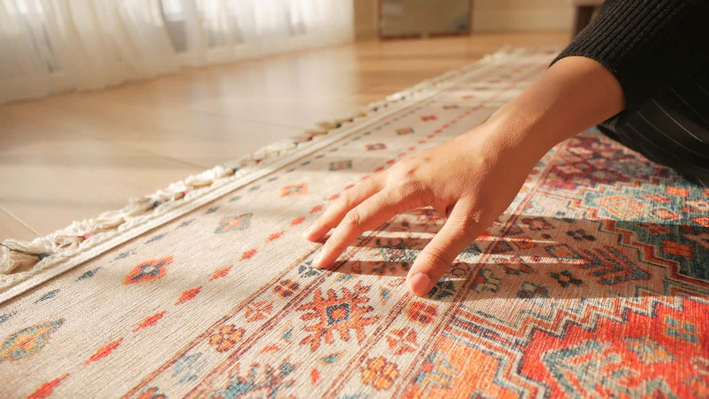 A hand gently touches a colorful, patterned rug on a wooden floor in bright sunlight.