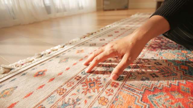 A hand gently touches a colorful, patterned rug on a wooden floor in bright sunlight.