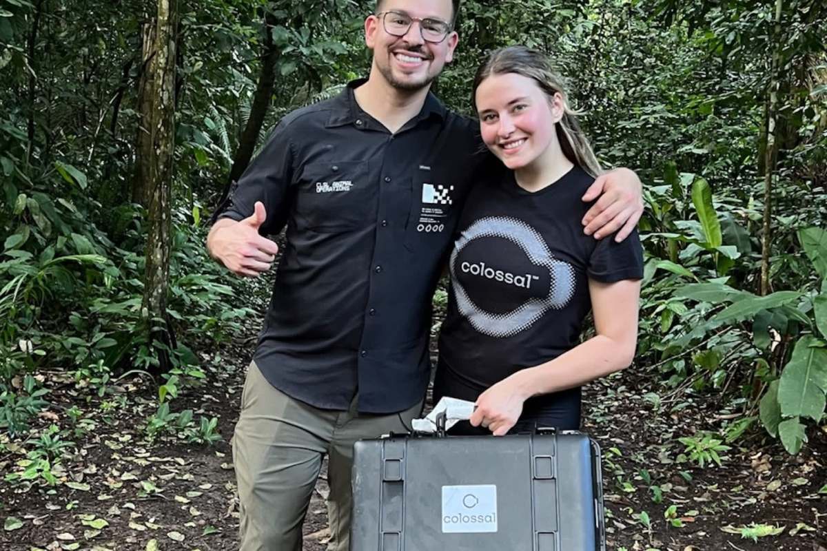 Two people stand in a lush forest, smiling and posing with a black case marked "Colossal."