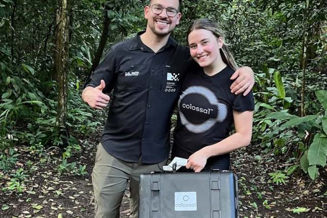 Two people stand in a lush forest, smiling and posing with a black case marked "Colossal."