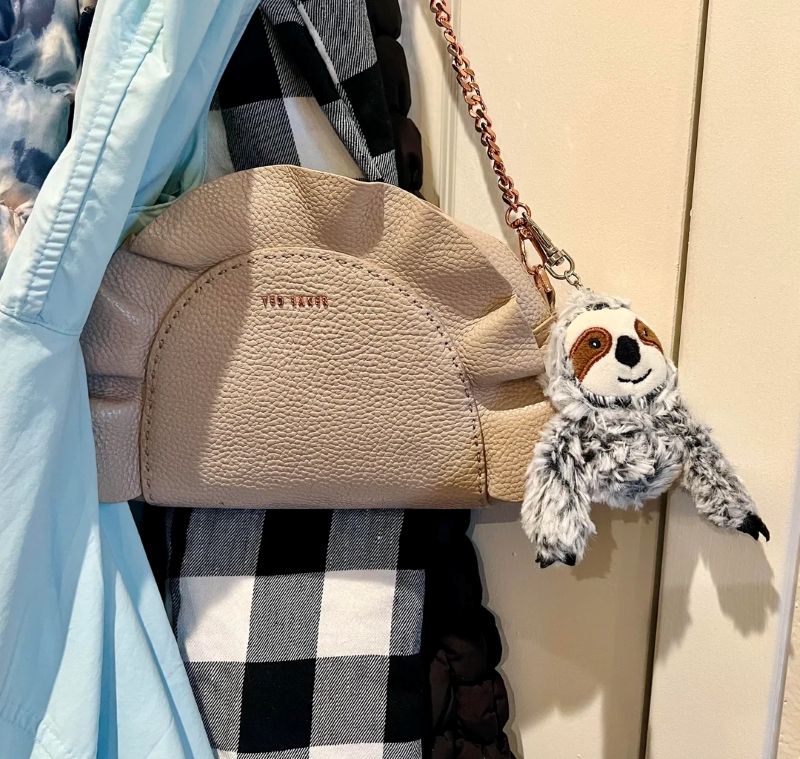 A stitched sloth plush toy attached as a charm to a beige leather purse. 
