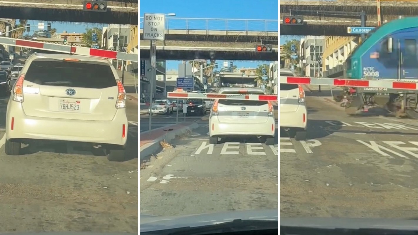 A white car waits at a lowered crossing gate with a train approaching in the background.