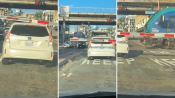 A white car waits at a lowered crossing gate with a train approaching in the background.