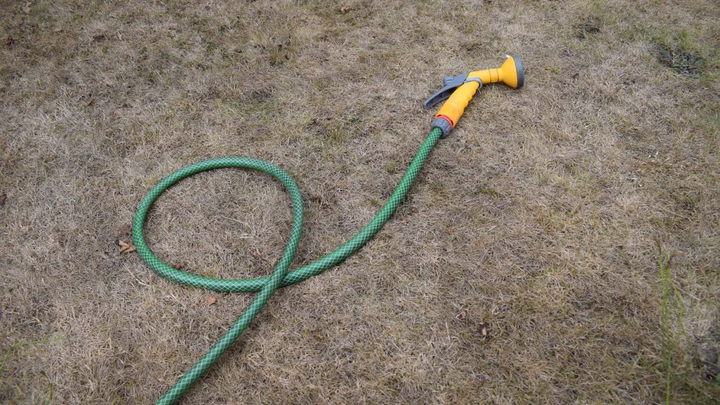 A green garden hose lies on dry, brown grass, with a yellow spray nozzle attached.