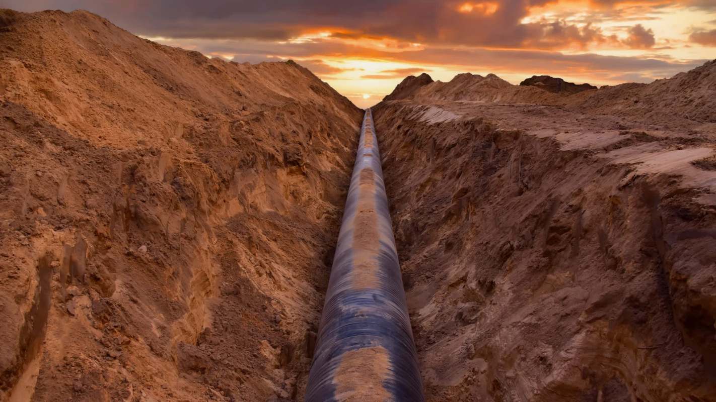 A long black pipeline is surrounded by freshly dug earth under a colorful sunset sky.