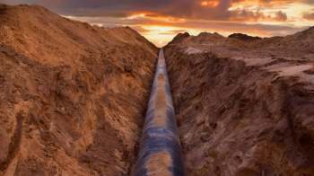 A long black pipeline is surrounded by freshly dug earth under a colorful sunset sky.