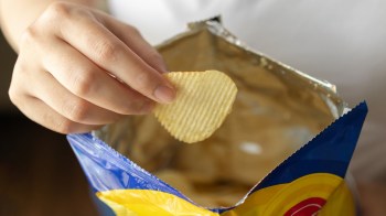 A hand is reaching into an open bag of potato chips, holding a single ridged chip.