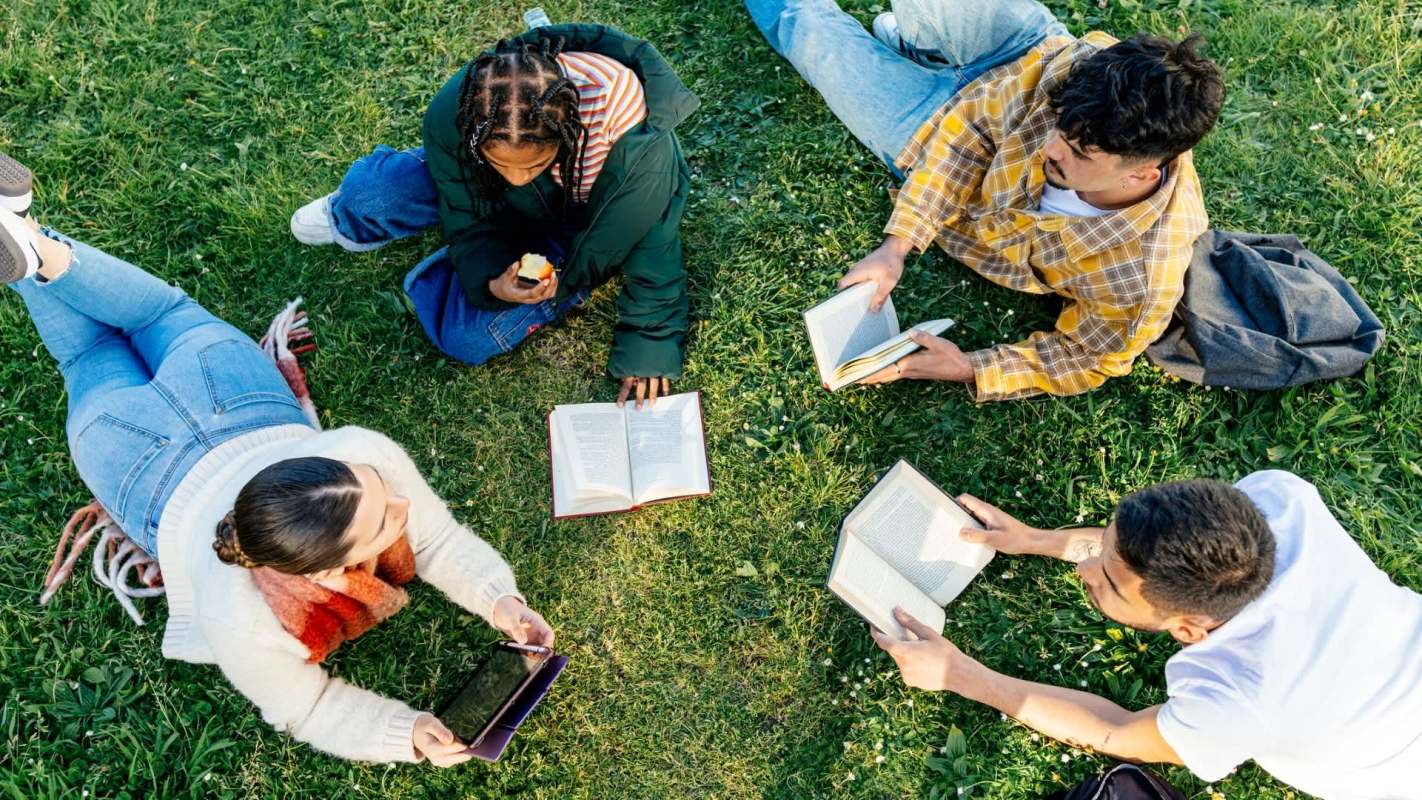 Four people are lounging on grass, reading books and using a tablet.