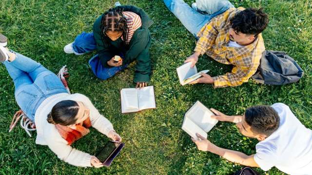 Four people are lounging on grass, reading books and using a tablet.