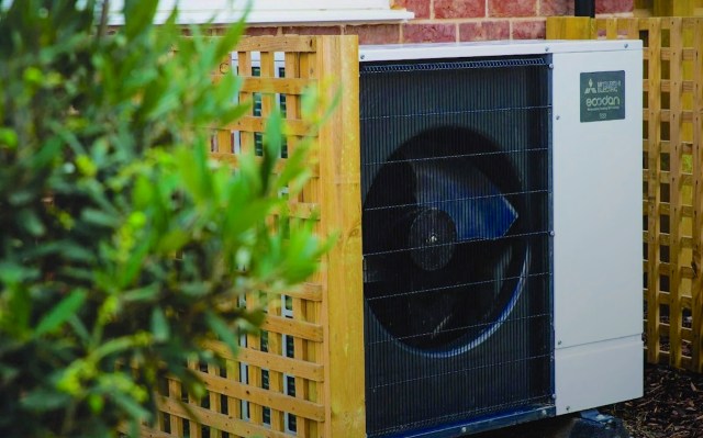 A Mitsubishi Ecodan outdoor heat pump unit partially obscured by greenery and a wooden lattice fence.