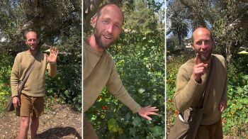 A man in a tan sweater and shorts poses in a garden, gesturing amid lush greenery and flowers.