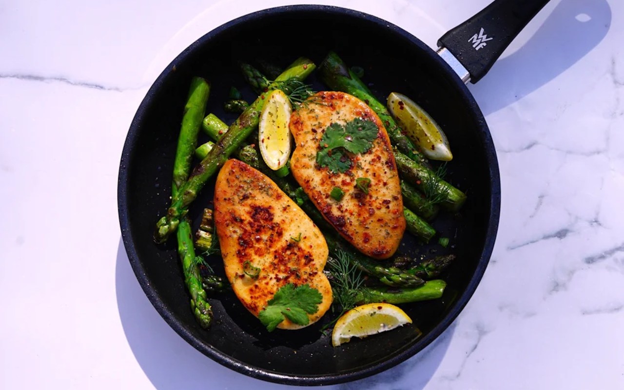 A skillet with two fillets, surrounded by asparagus and garnished with lemon slices and fresh herbs.
