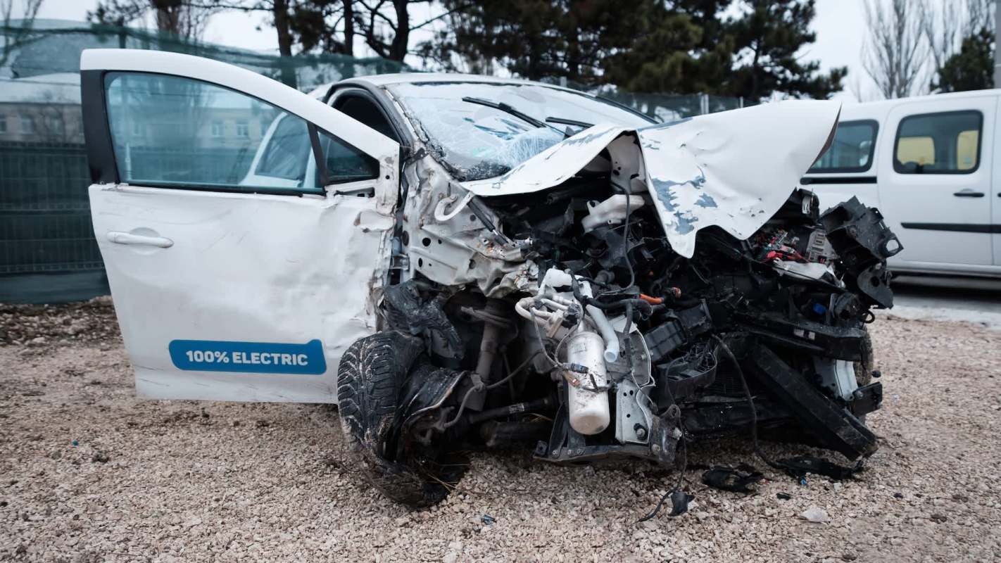 A heavily damaged electric car with a crushed front and broken windshield parked on gravel.