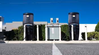 Two electric vehicle charging stations in front of green bushes.