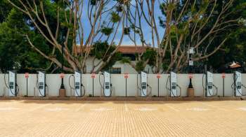 A row of electric vehicle charging stations under a canopy of trees in a paved area.