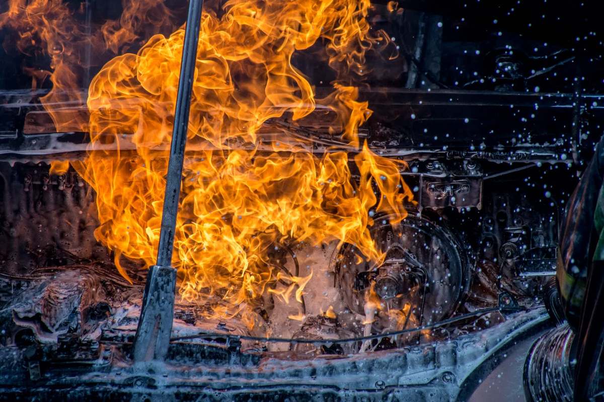 Flames engulf a car engine component, as water and foam attempt to extinguish the fire.