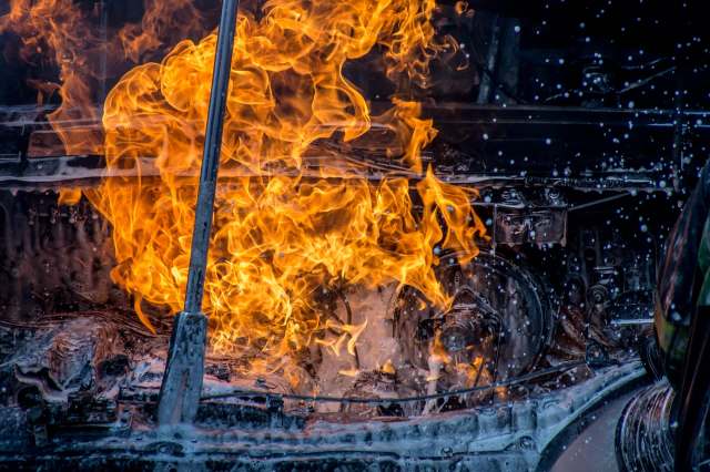 Flames engulf a car engine component, as water and foam attempt to extinguish the fire.