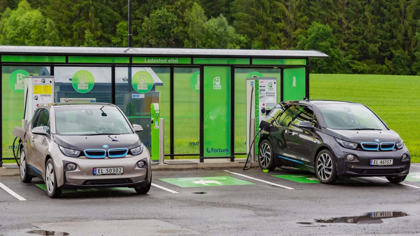 Two electric cars parked at charging stations near a modern facility surrounded by greenery.