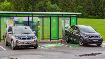 Two electric cars parked at charging stations near a modern facility surrounded by greenery.