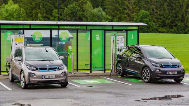 Two electric cars parked at charging stations near a modern facility surrounded by greenery.