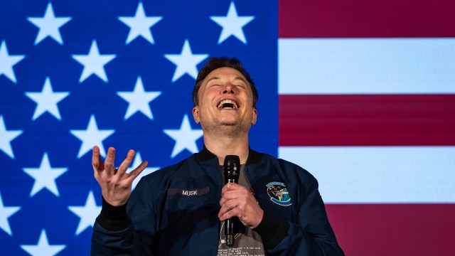 Elon Musk wearing a blue jacket smiling and speaking into a microphone against a background of stars and stripes.