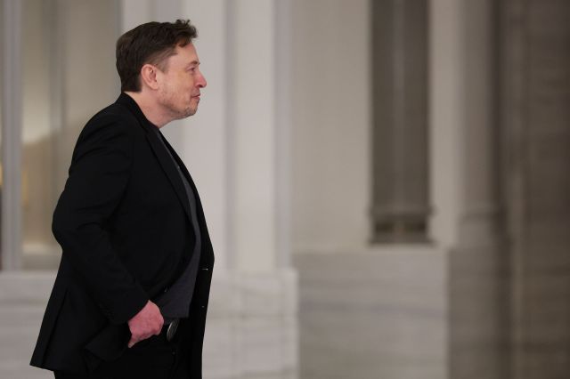 Elon Musk in a black jacket, walking with his hands in his pockets.